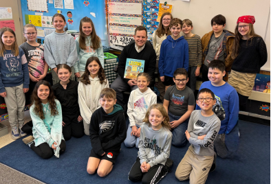  students seated on the carpet around Dr. Yanni seated in a chair holding the book he read